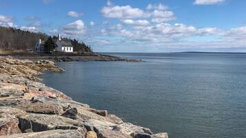 A rocky shoreline with a white building on a small island, surrounded by calm water and a clear sky.
