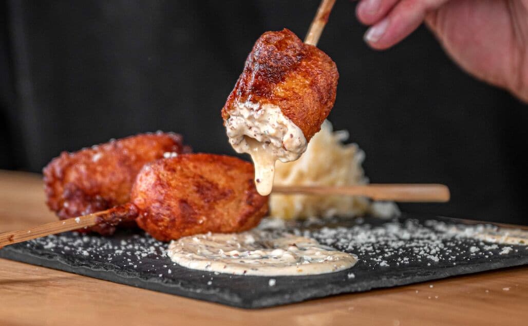 Una mano sostiene un pincho con un bocado de comida, posiblemente una albóndiga, sobre un plato de pizarra negra con un objeto redondo pequeño y queso rallado.