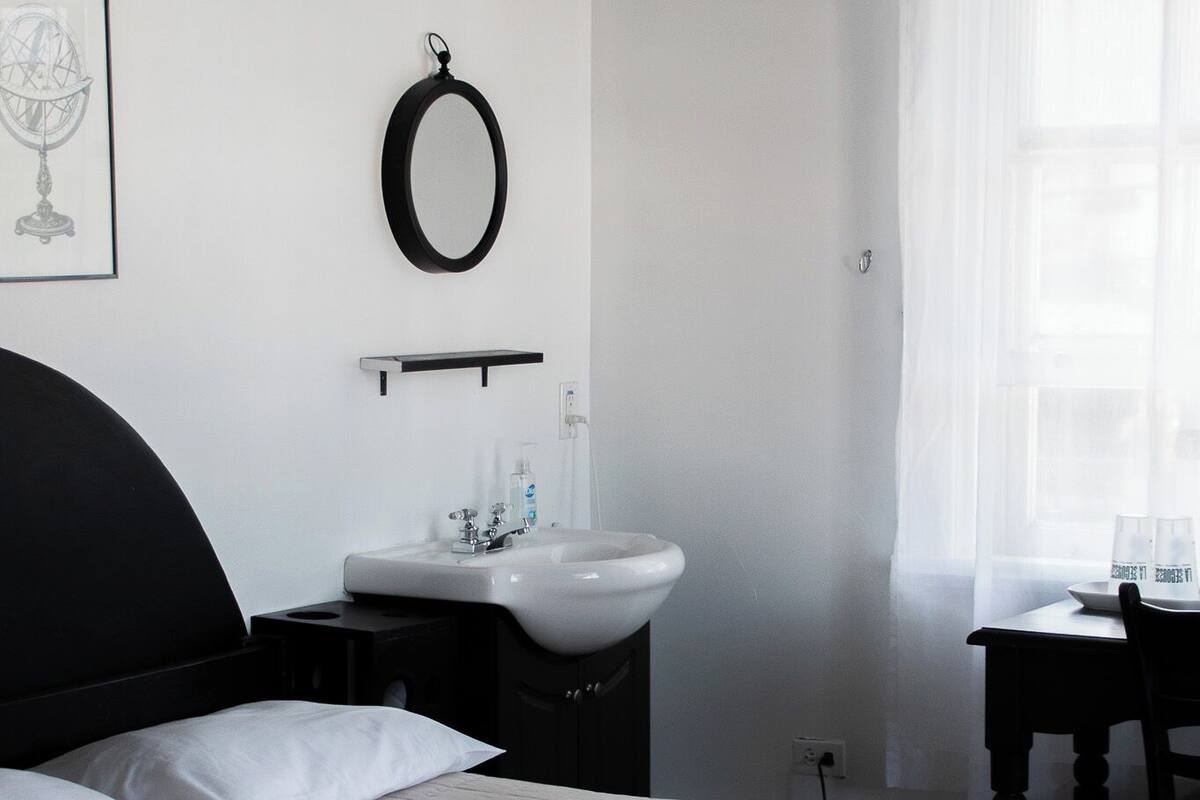 Habitación de hotel con cama, mesita de noche, espejo, lavamanos y ventana con cortinas traslúcidas.