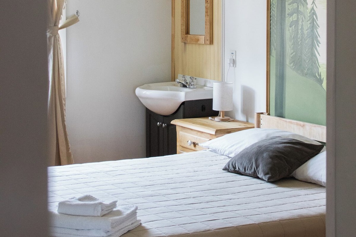 Un dormitorio con cama, mesita de noche con lámpara, espejo y ventana con cortinas.