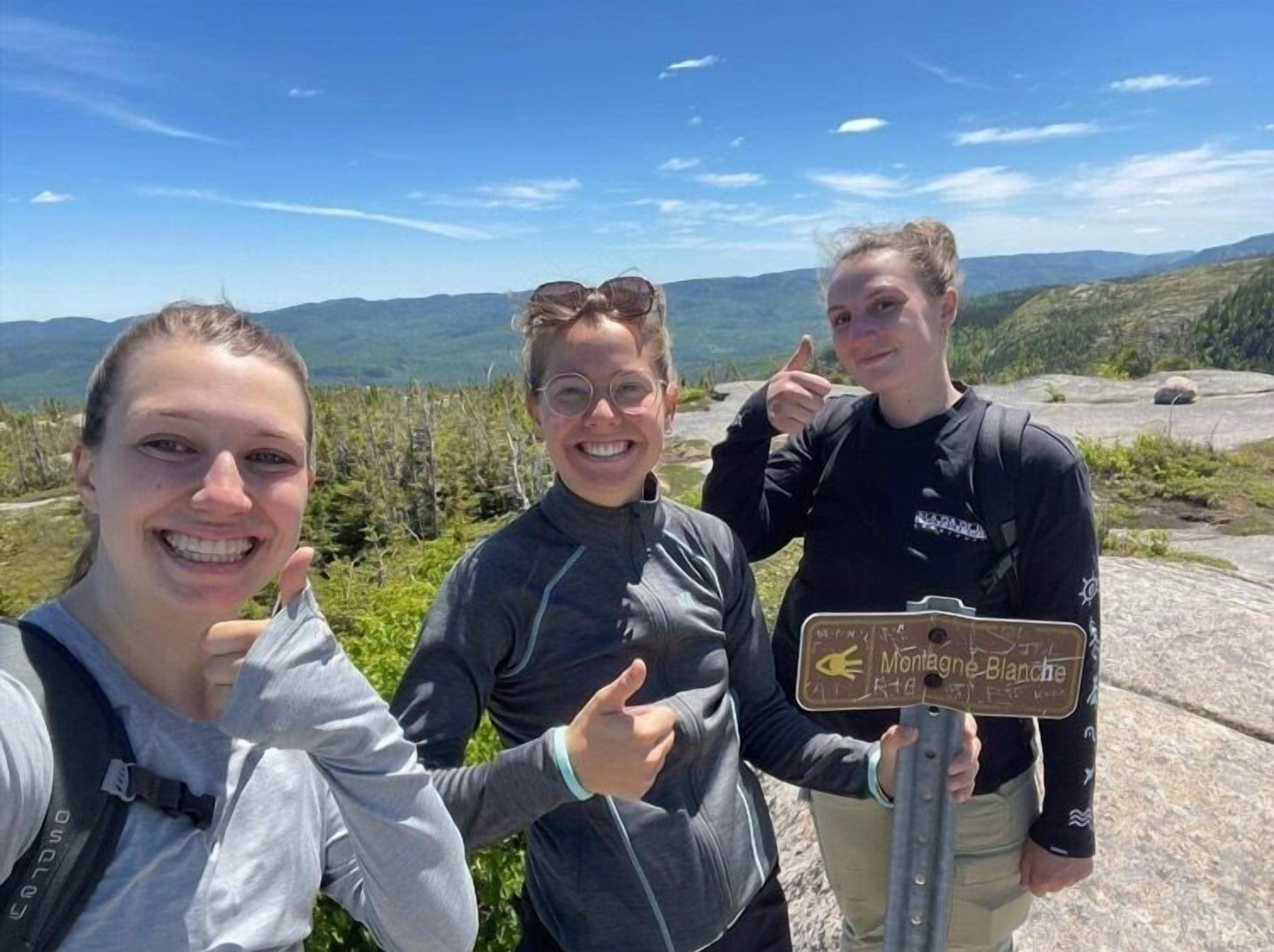 Three people standing near a signpost with a view of mountains in the background.