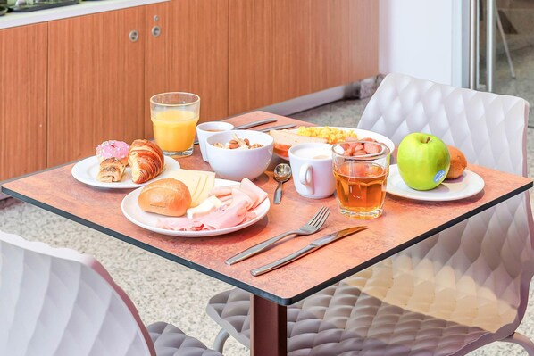 A breakfast table with croissants, a plate of ham and cheese, a glass of orange juice, a bowl of cereal, a cup of coffee, a glass of tea, and an apple.