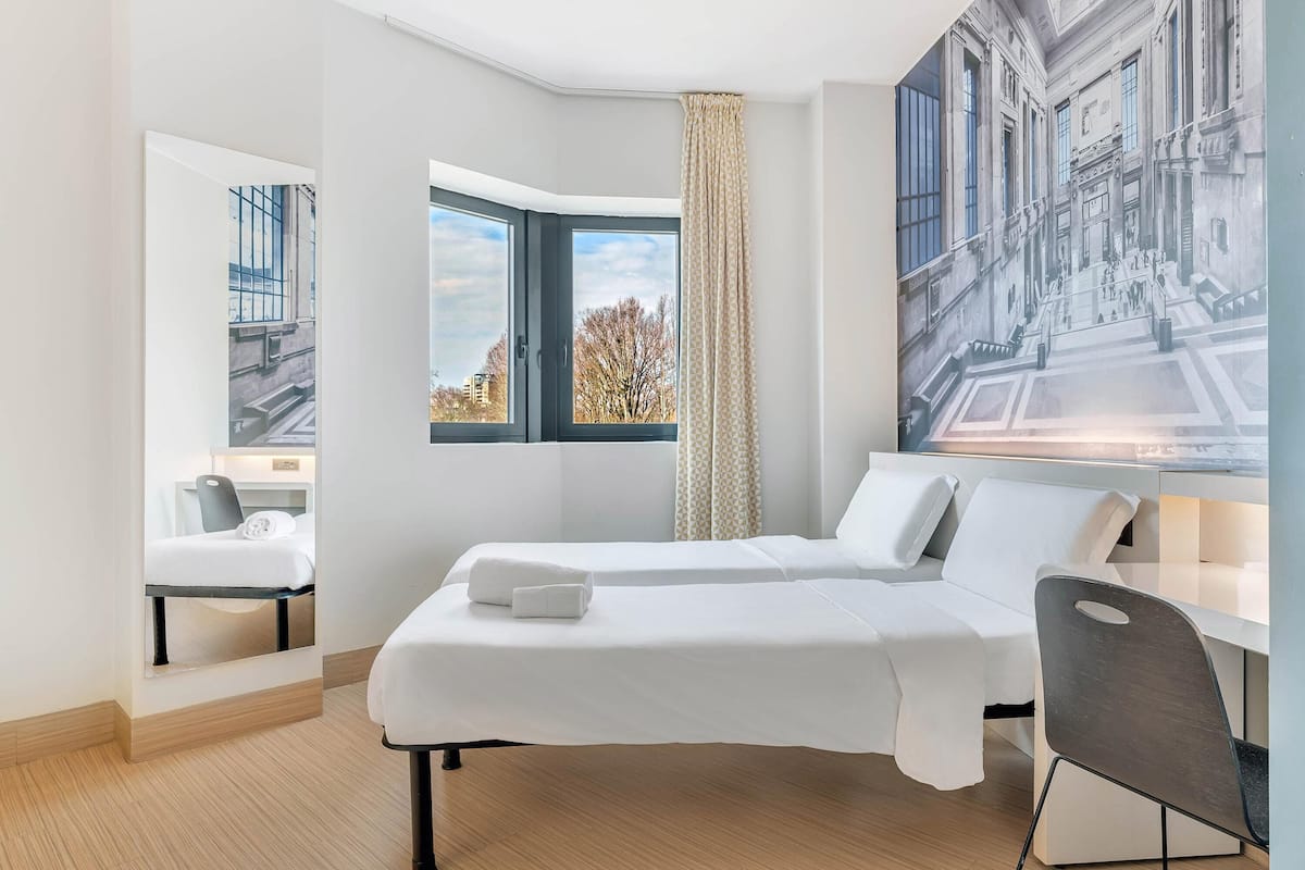 Une chambre d’hôtel moderne dotée d’un grand lit, d’un bureau, d’une chaise, d’une fenêtre donnant sur des immeubles et d’un miroir qui reflète la pièce.