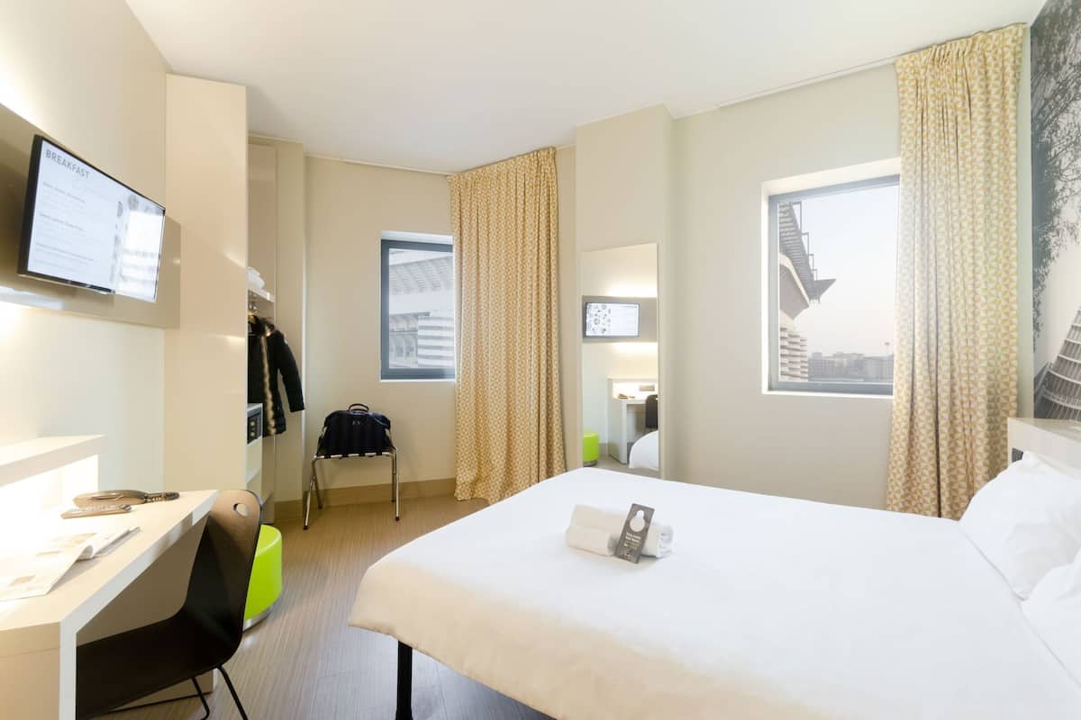 Une chambre d’hôtel avec un lit, un bureau, une chaise et un téléviseur fixé au mur.