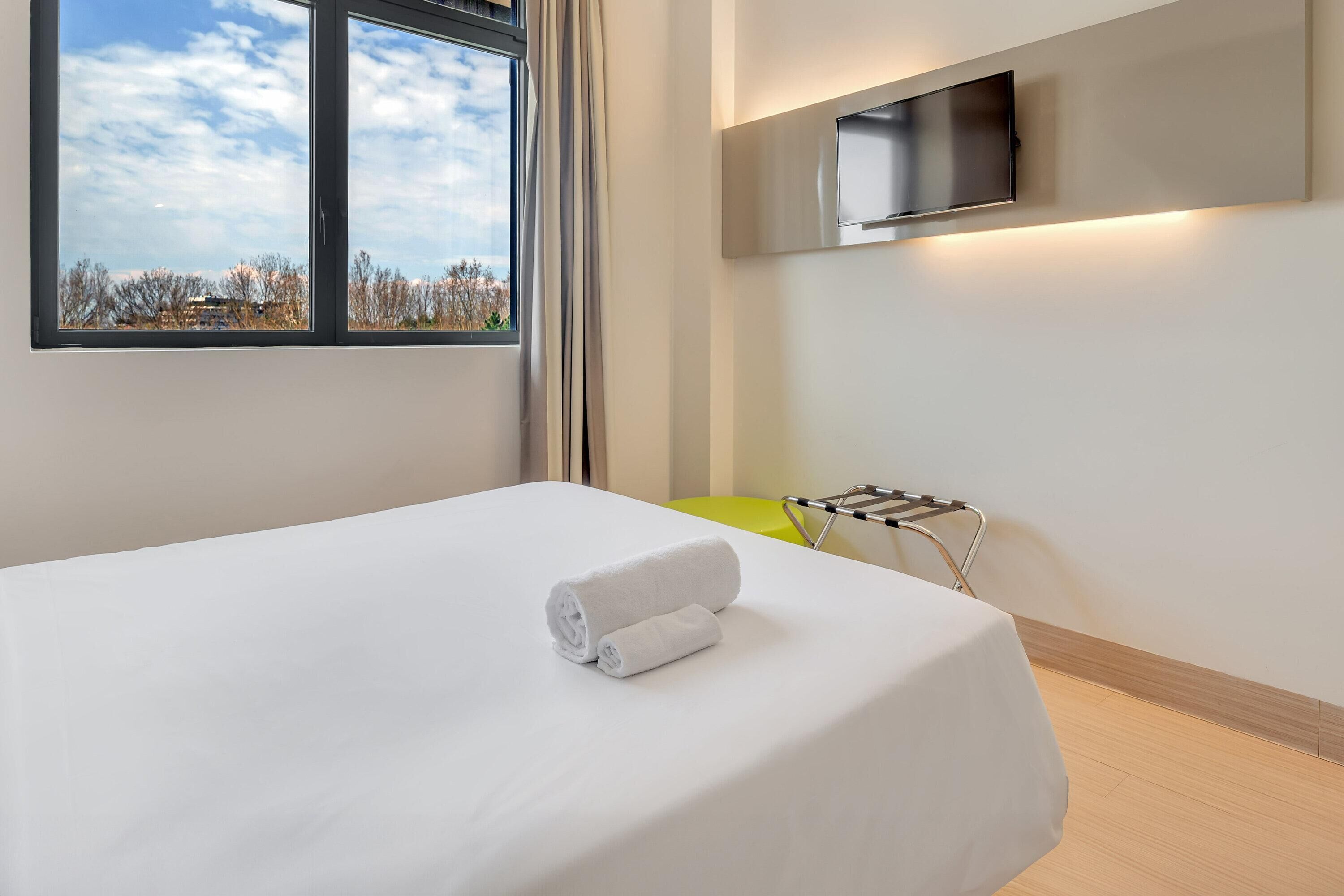 Une chambre d’hôtel avec un lit, un téléviseur fixé au mur, une fenêtre donnant sur des arbres et une serviette sur le lit.