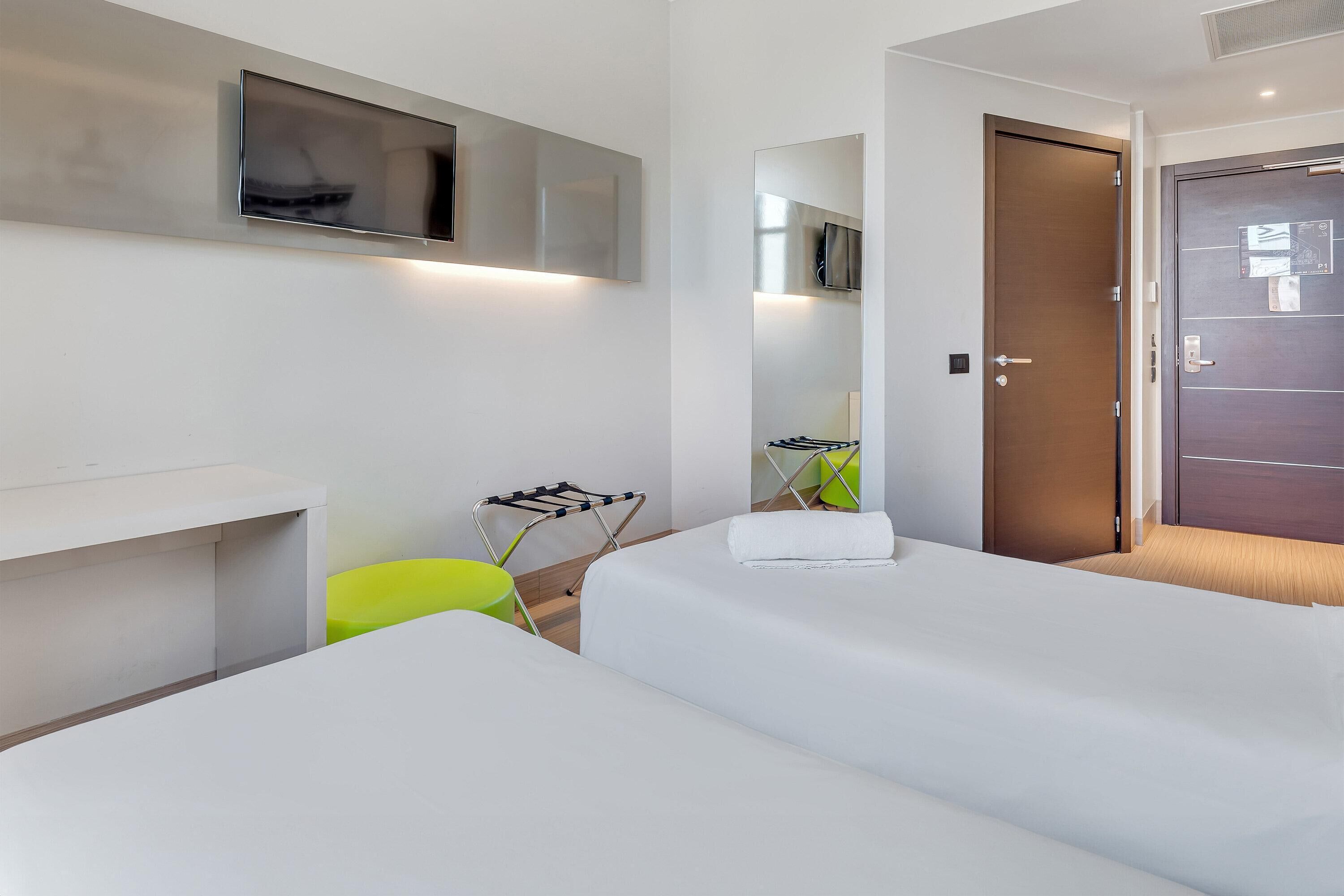 A hotel room with two beds, a TV mounted on the wall, a door with a handle, and a small table with a chair.