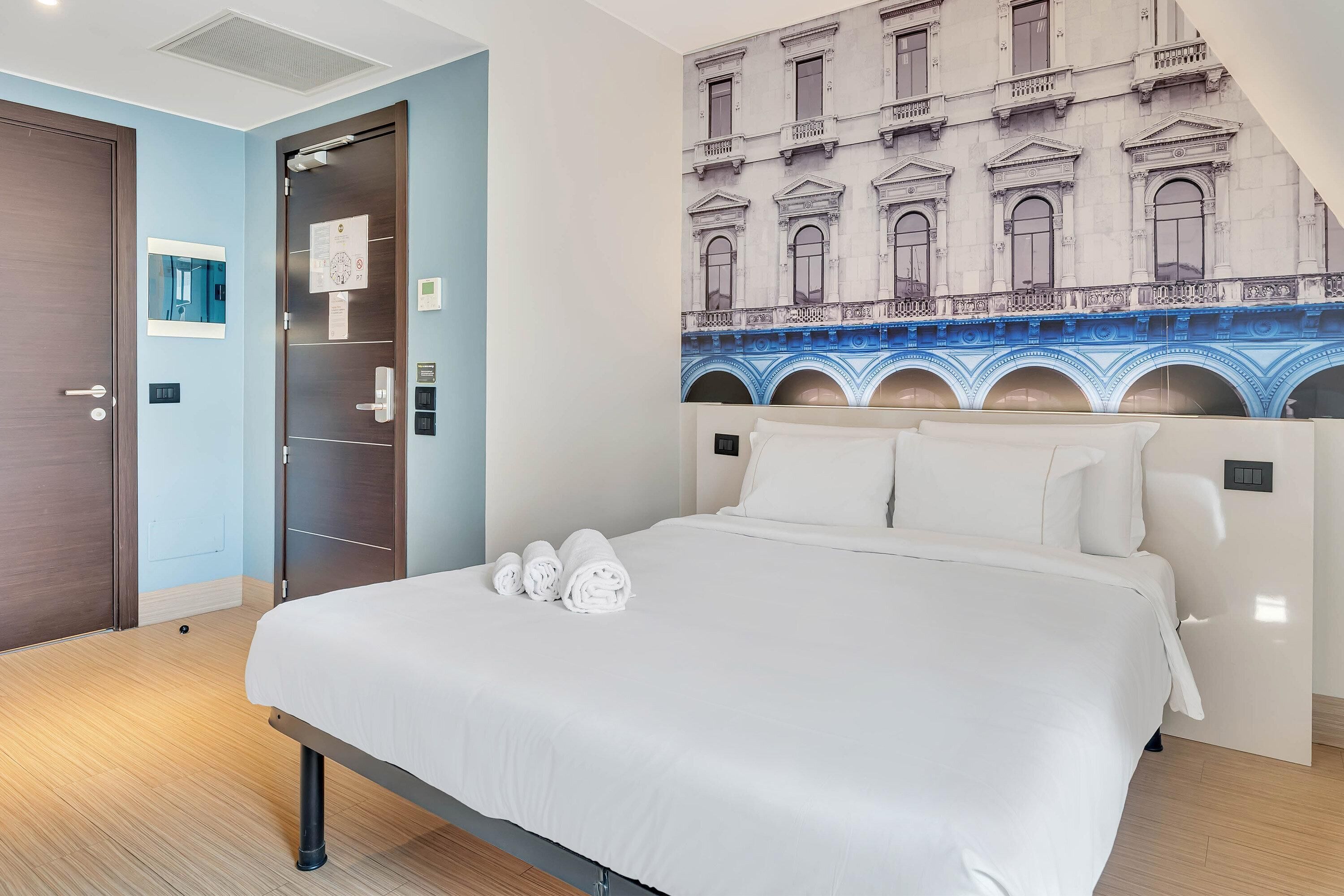 A hotel room with a bed, a door, a wall with a mural, and a light switch.