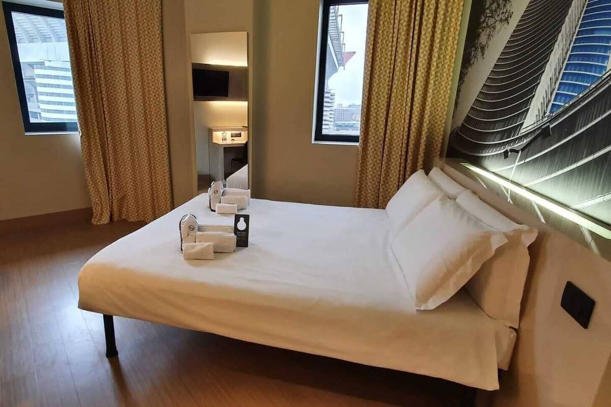 Une chambre d’hôtel avec un lit, un bureau, une chaise, une poubelle verte et une fenêtre avec des rideaux.