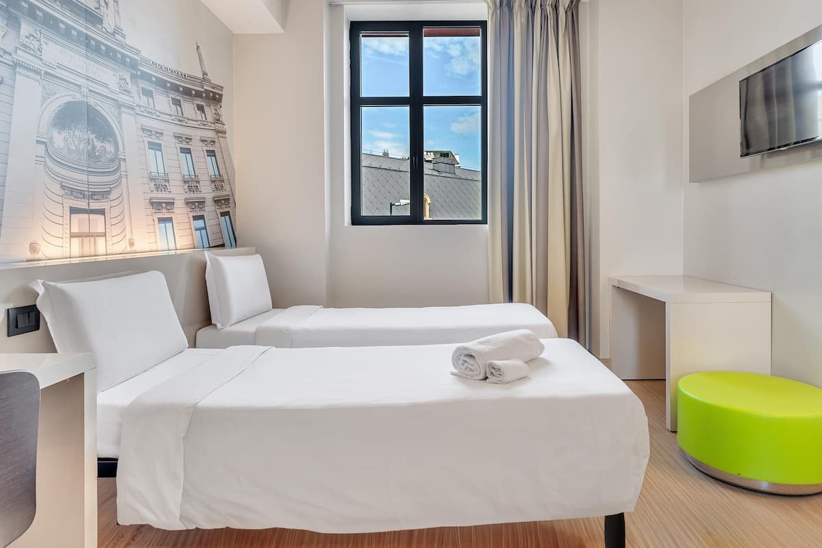 Une chambre d’hôtel avec deux lits, une grande fenêtre donnant sur un beau paysage, un téléviseur à écran plat et un pouf vert.