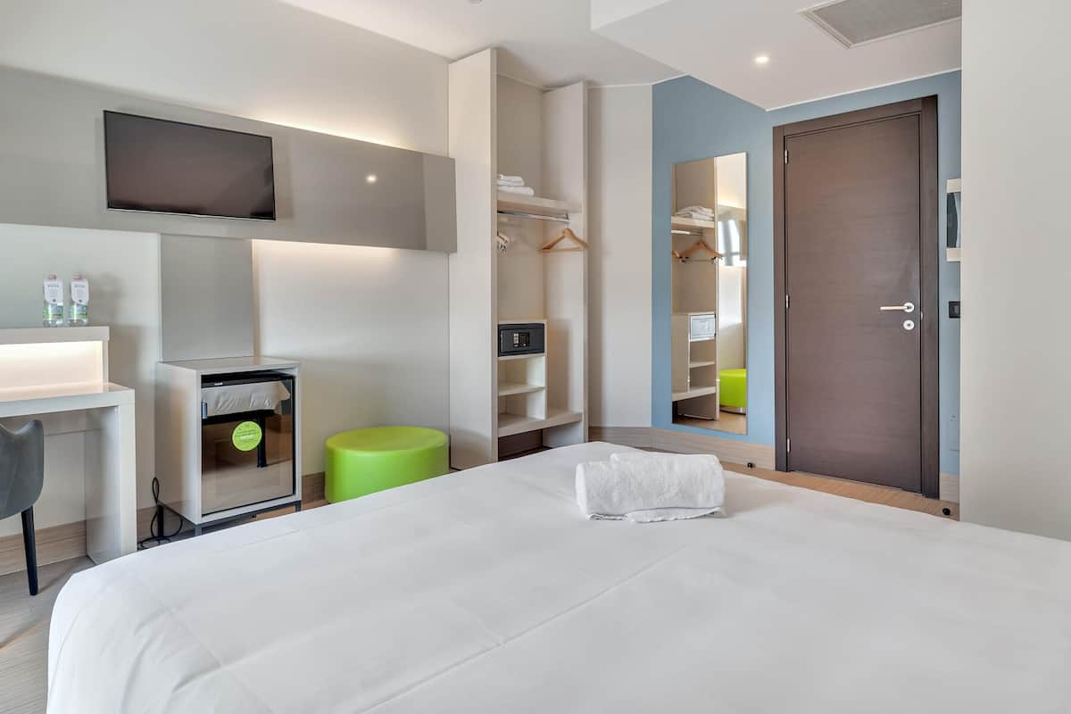 Une chambre d’hôtel moderne dotée d’un grand lit, d’un bureau avec une chaise, d’un téléviseur fixé au mur, d’une armoire et d’une salle de bain avec des toilettes et un lavabo.