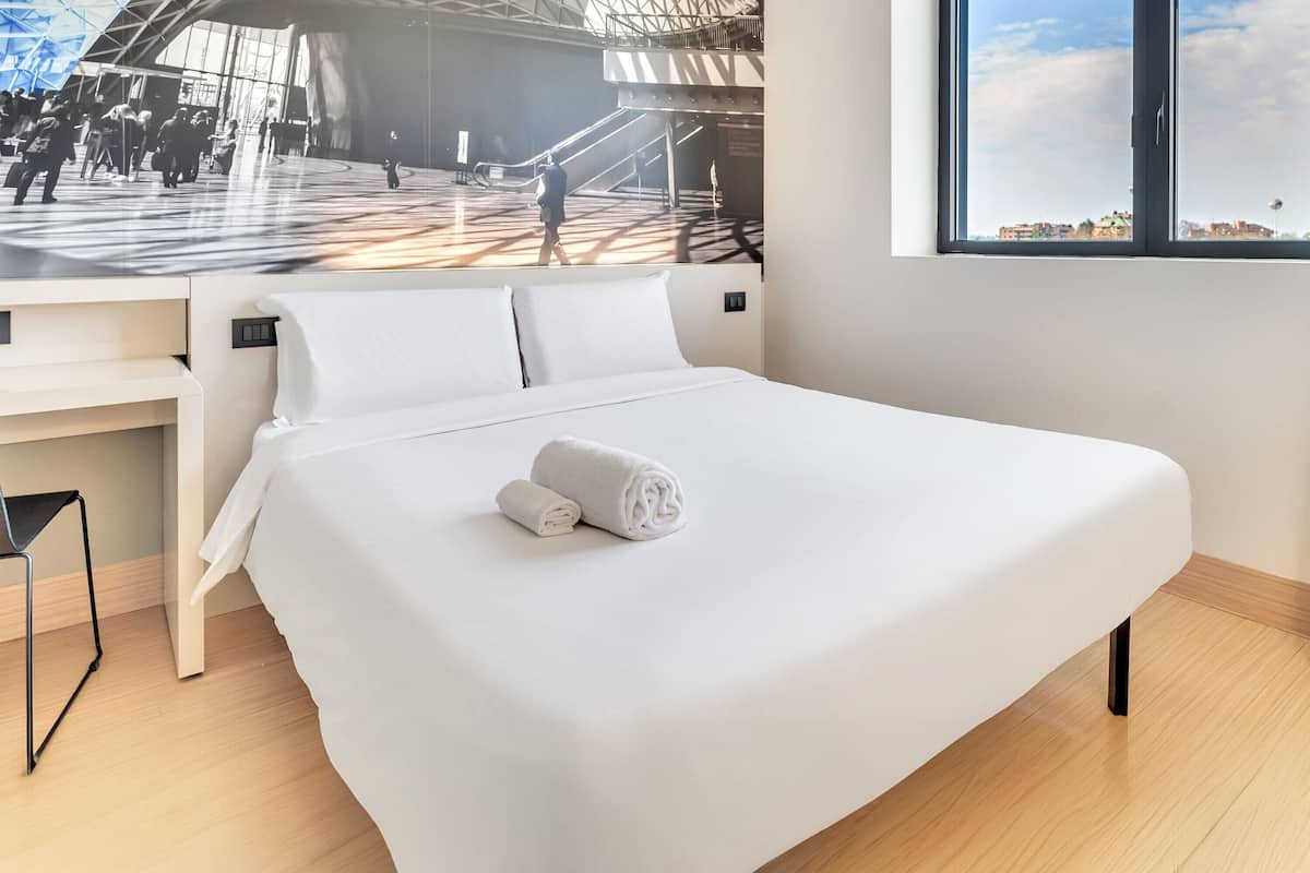 Une chambre d’hôtel moderne, dotée d’un grand lit, d’un bureau et offrant une vue sur un espace public animé grâce à une grande fenêtre.