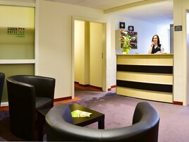 Zona de recepción de un hotel, con mostrador de recepción, área de espera con sillas y piso alfombrado.