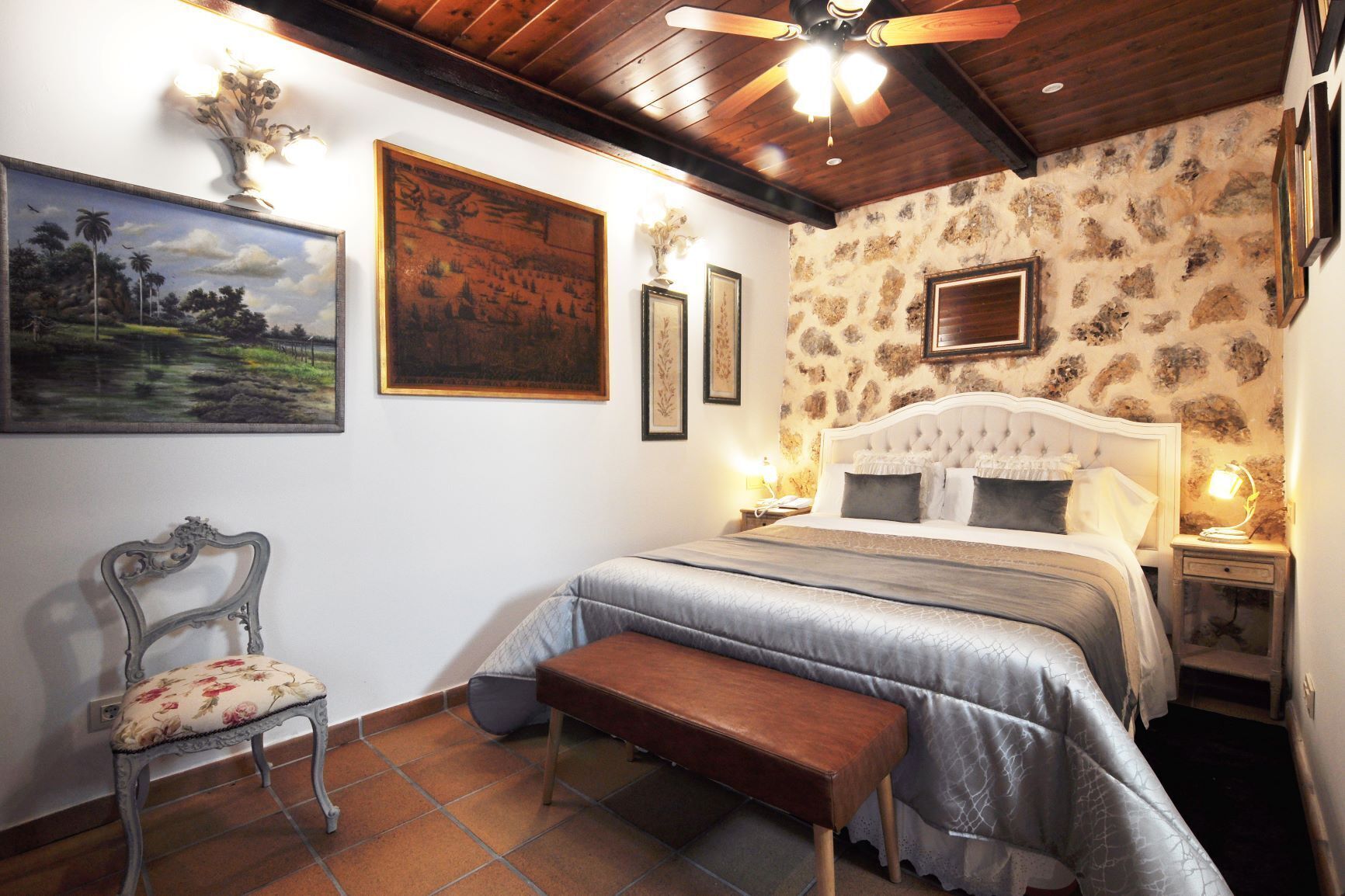 A bedroom with a large bed, a chair, a bench, and framed artwork on the walls.