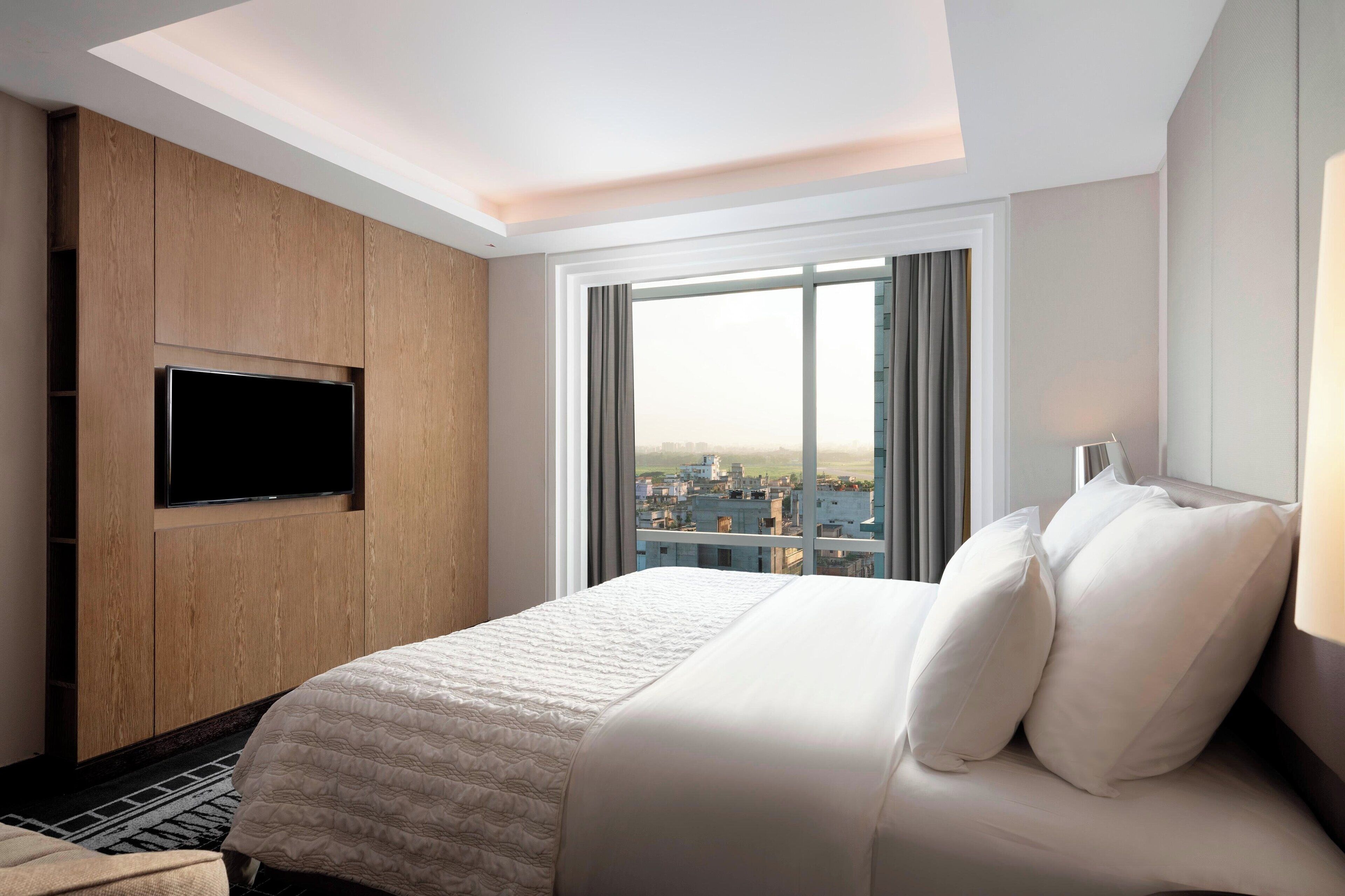 Une chambre d’hôtel dotée d’un grand lit, d’un téléviseur à écran plat fixé sur un panneau en bois et offrant une vue sur la ville grâce à de grandes fenêtres.