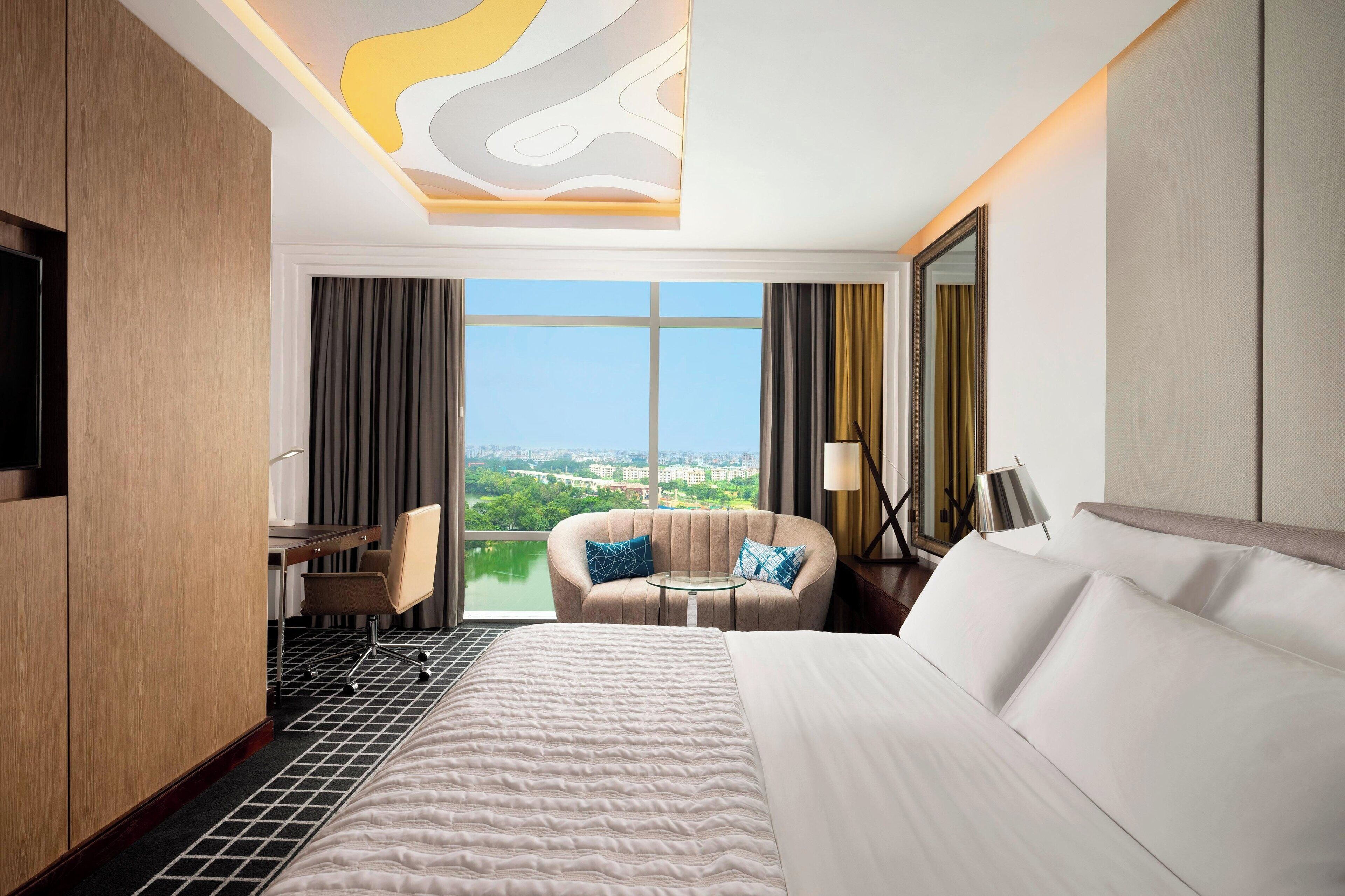A hotel room with a large bed, a desk with a chair, a sofa, and a view of a cityscape through large windows.