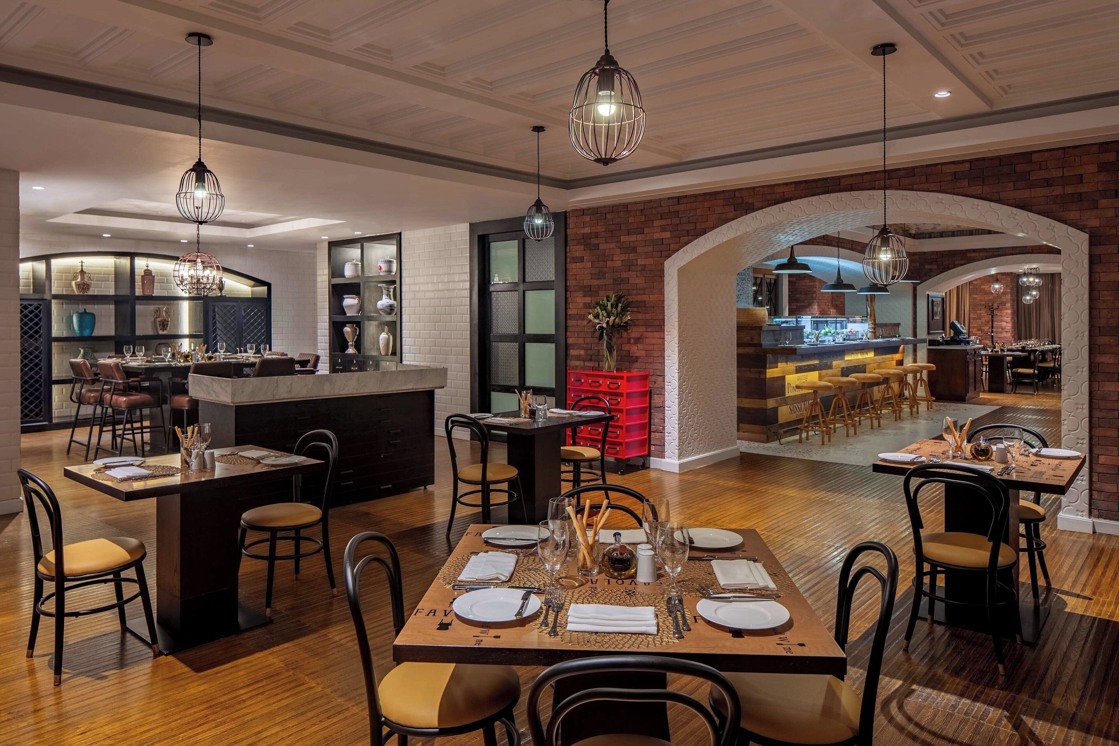 Un comedor espacioso con varias mesas dispuestas para los huéspedes, una cocina con electrodomésticos modernos y una pared de ladrillo con un arco de entrada a otra zona de comedor.