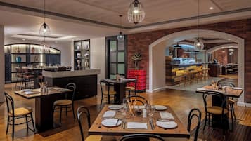 Un comedor espacioso con varias mesas dispuestas para los huéspedes, una cocina con electrodomésticos modernos y una pared de ladrillo con un arco de entrada a otra zona de comedor.