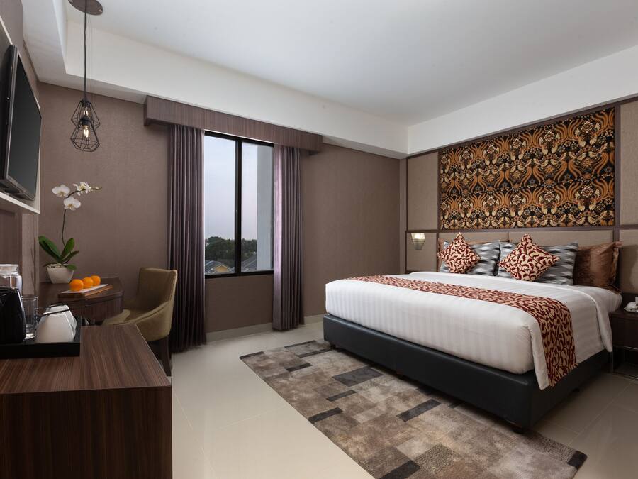 A modern hotel room with a large bed, a desk with a coffee maker, a TV, and a large window with curtains.