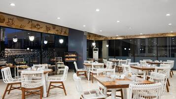 Un restaurante espacioso con sillas de mimbre blanco y mesas de madera, un ventanal grande y una pared con diseños decorativos.