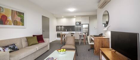 A modern living room with a sofa, coffee table, and TV. There is a kitchen area with a fridge, microwave, and dining table.