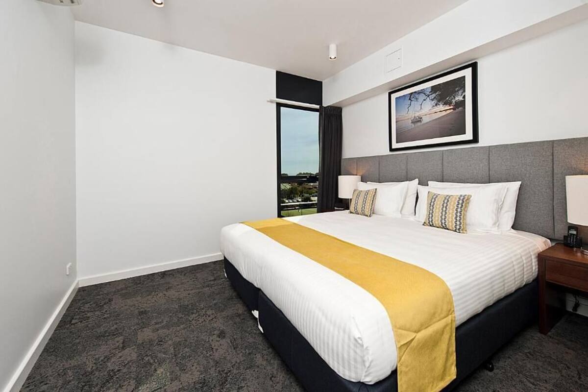 A hotel room with a large bed, a view of the outdoors, a framed picture on the wall, and a nightstand with a lamp.