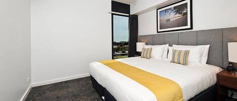 A hotel room with a large bed, a view of the outdoors, a framed picture on the wall, and a nightstand with a lamp.