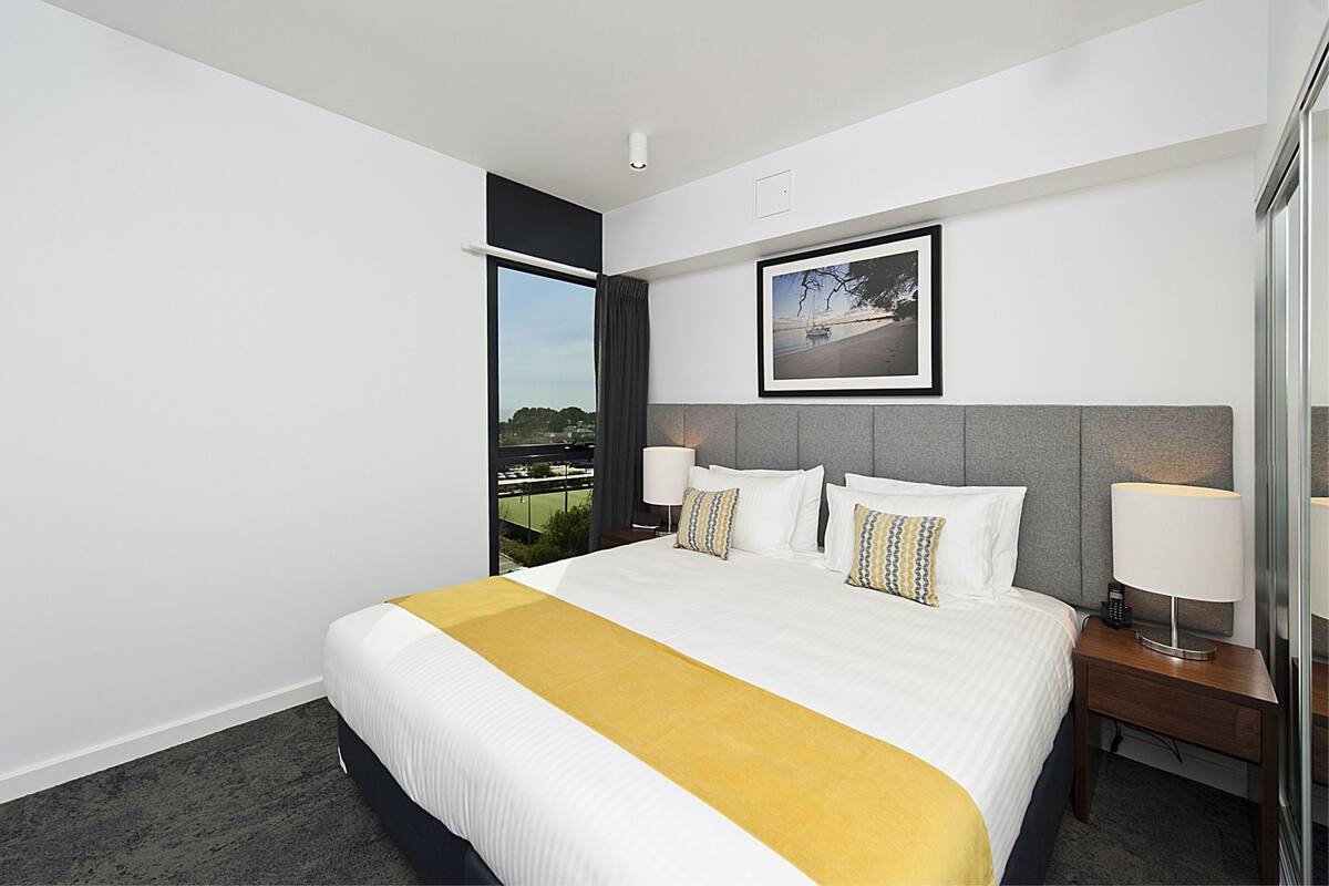 A hotel room with a large bed, a bedside table, a lamp, a framed picture on the wall, and a window with a view.
