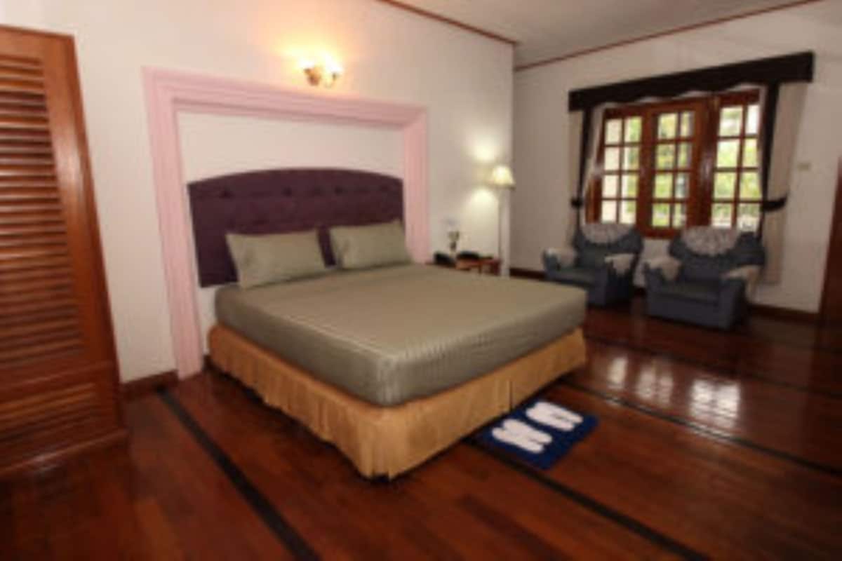 A bedroom with a large bed, wooden flooring, a chair, and a window with blinds.
