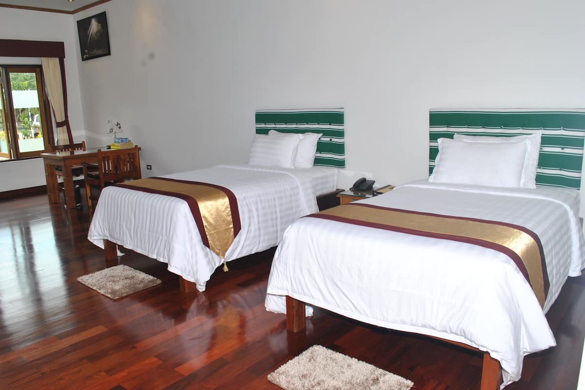 A hotel room with two beds, wooden flooring, a small table with chairs, and a window with curtains.