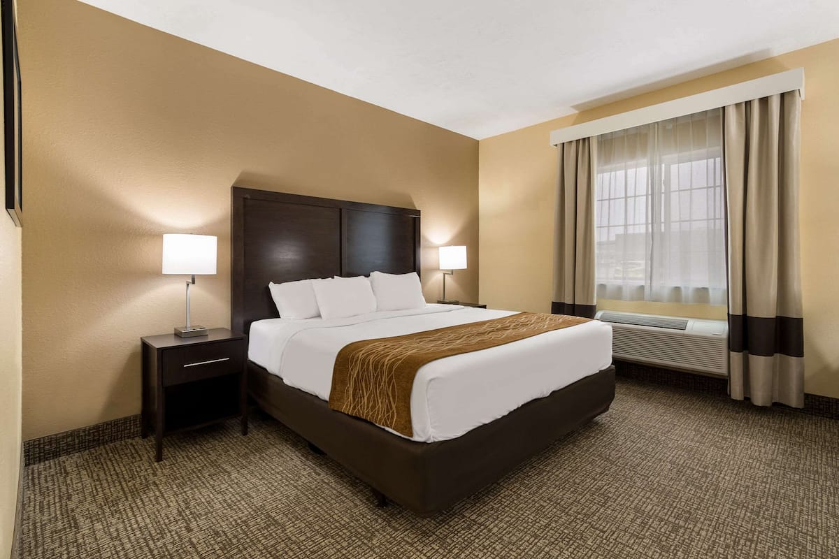 A hotel room with a large bed, two bedside tables with lamps, a window with curtains, and a wall-mounted air conditioning unit.