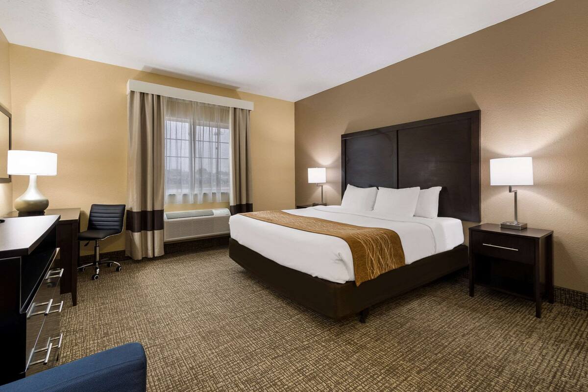 A hotel room with a large bed, a desk, a chair, a nightstand, and a window with curtains.