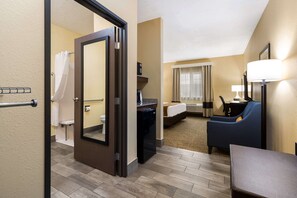 Habitación de hotel con cama, escritorio, silla, lámpara y baño visible a través de una puerta abierta.