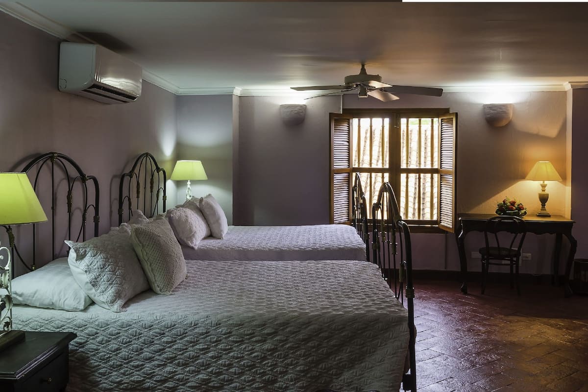 Un dormitorio con dos camas, un ventilador de techo, una ventana con persianas y un escritorio con una lámpara.