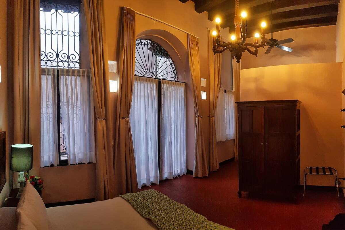 Una habitación de hotel con cama, un armario de madera, una araña y una ventana con cortinas.