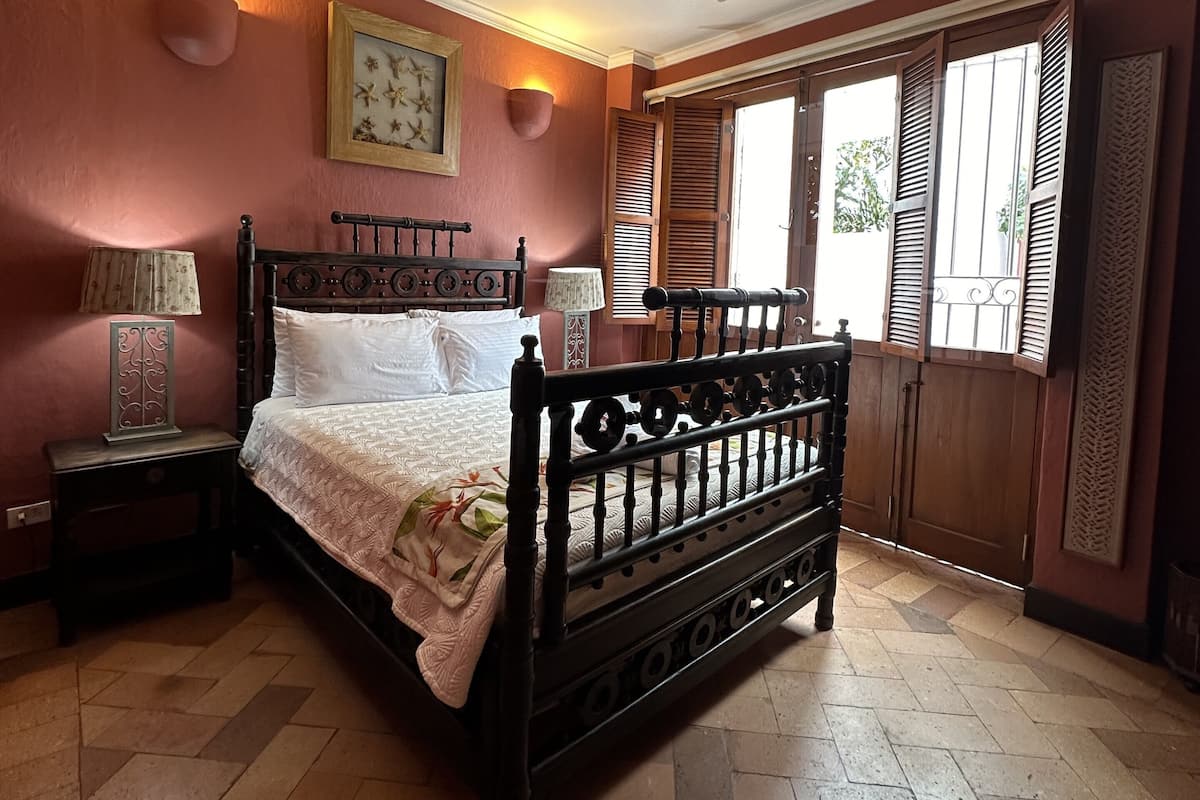 Un dormitorio con cama de madera, ventilador de techo, mesita de noche con lámpara, luz de pared y ventana con contraventanas de madera.