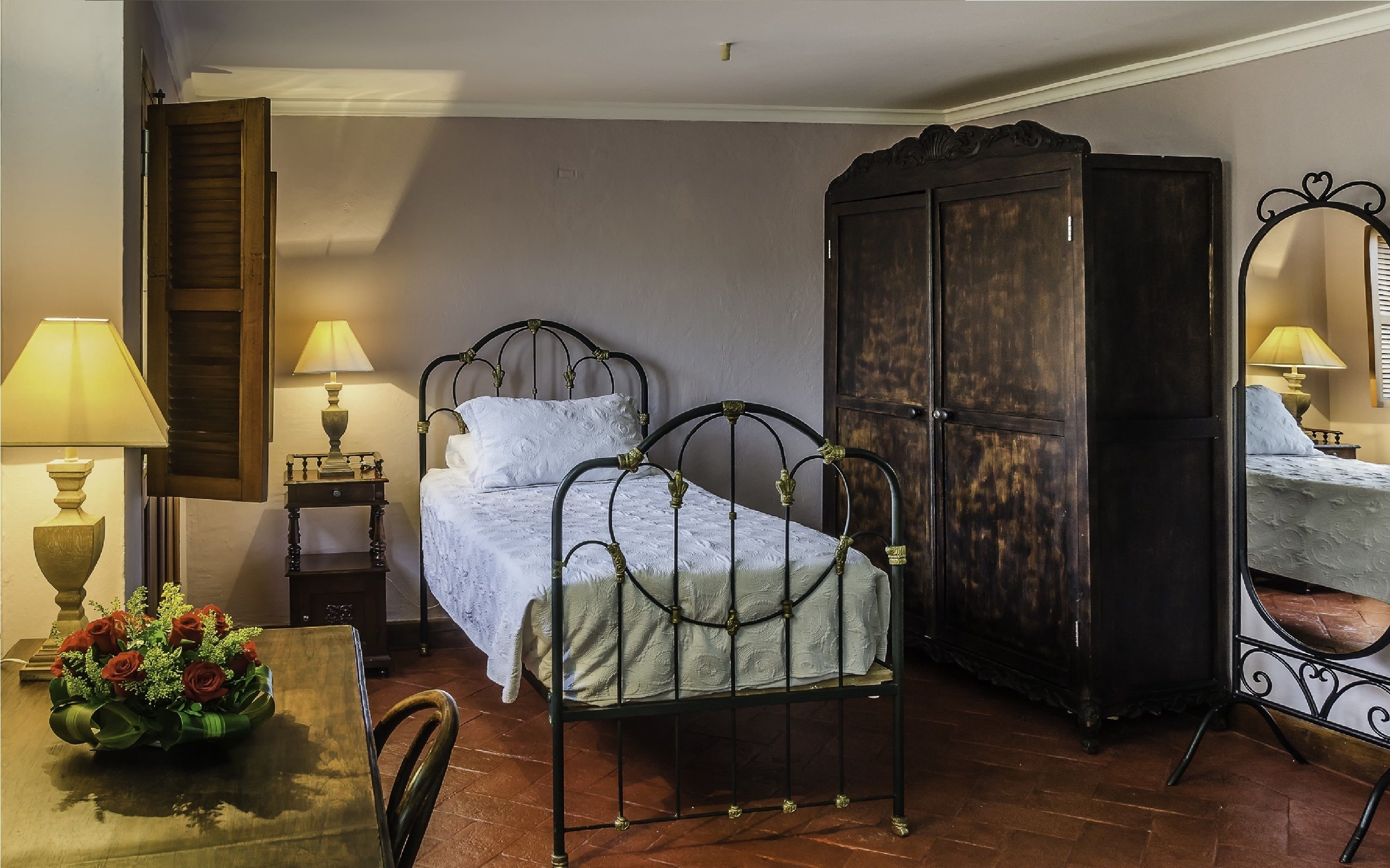 A room with a bed, a wooden wardrobe, a table with a lamp and flowers, and a chair.
