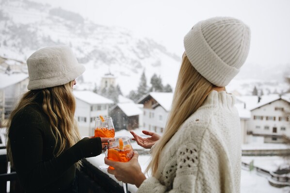 Dos personas sosteniendo vasos con bebidas, de pie junto a una ventana que da a un pueblo nevado con casas y montañas al fondo.