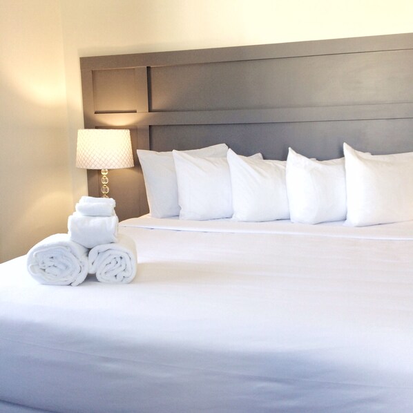 Una cama bien hecha con sábanas y almohadas blancas, una lámpara en la mesita de noche y toallas enrolladas.