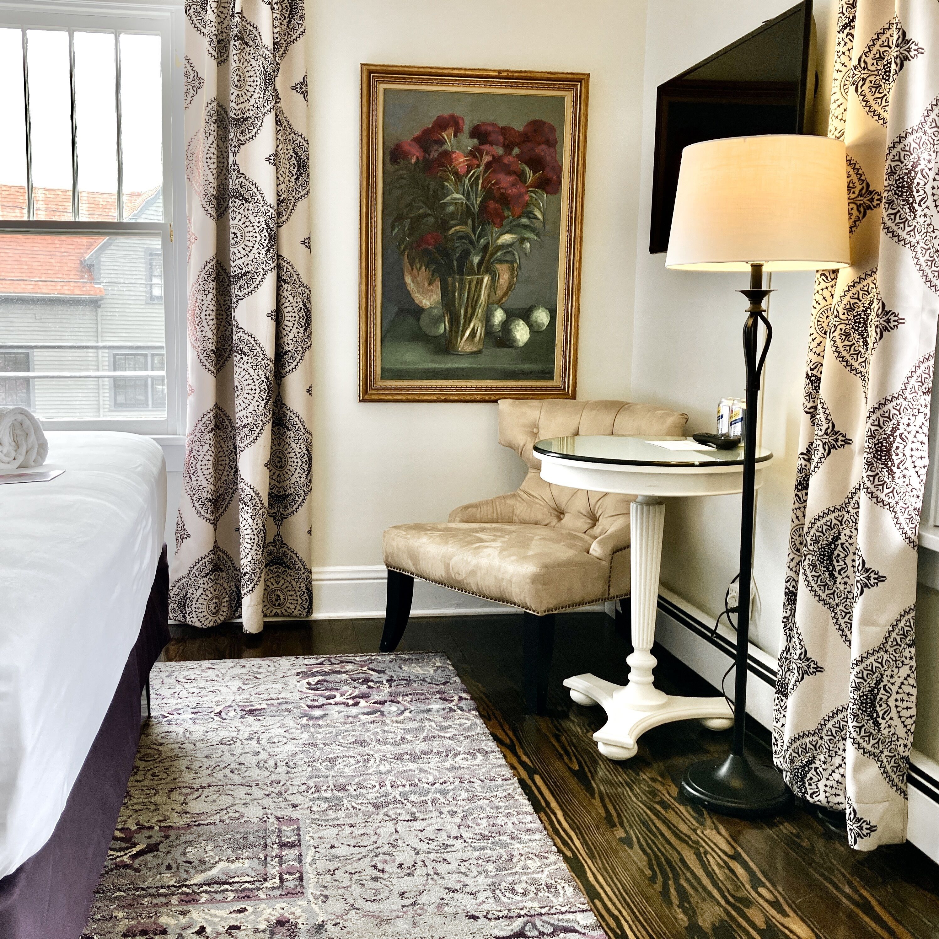 A hotel room with a bed, a round table with a lamp, a painting of flowers, and patterned curtains.