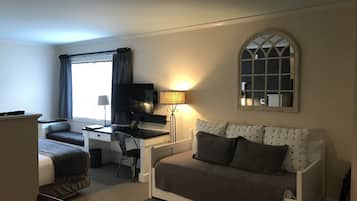 A compact hotel room with a bed, a sofa, a desk, a chair, a TV, and a window with curtains.
