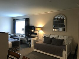 A compact hotel room with a bed, a sofa, a desk, a chair, a TV, and a window with curtains.