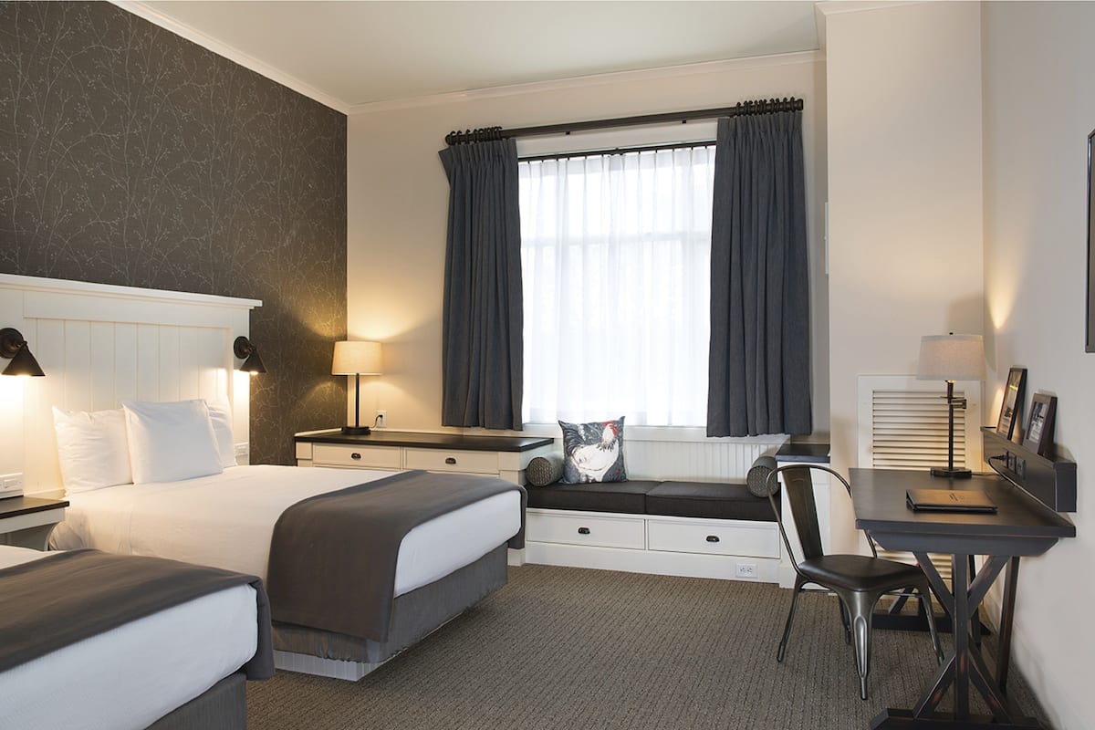 Una habitación de hotel con cama, escritorio, silla, ventana con cortinas y lámpara.