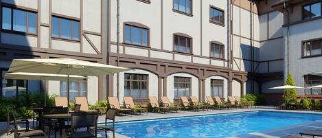 Un área de piscina con sillones de descanso y mesas, rodeada por un edificio con múltiples ventanales y suelo de baldosas.