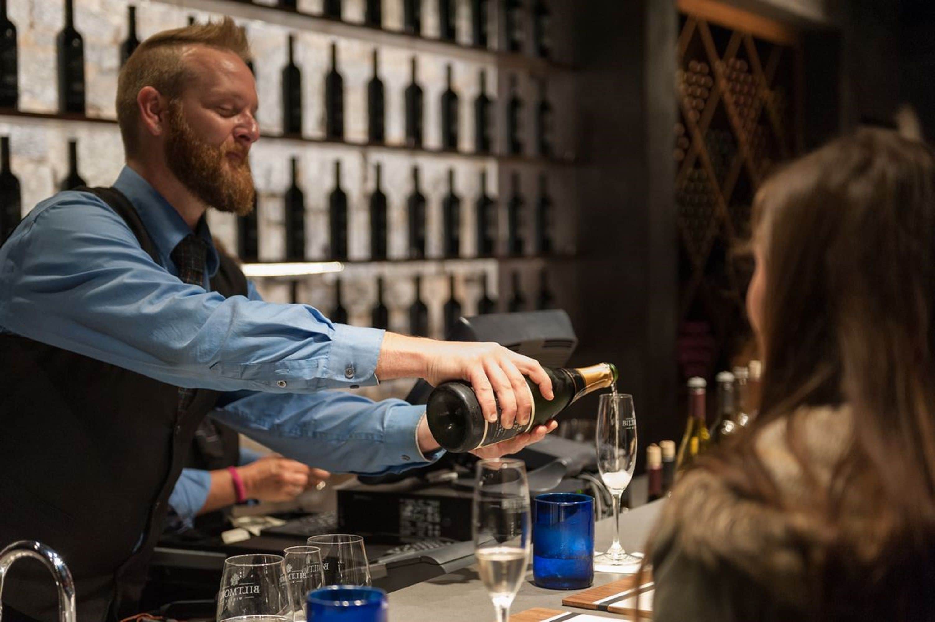 En bartender hælder drik i et glas bag en bar med en vinreol i baggrunden.