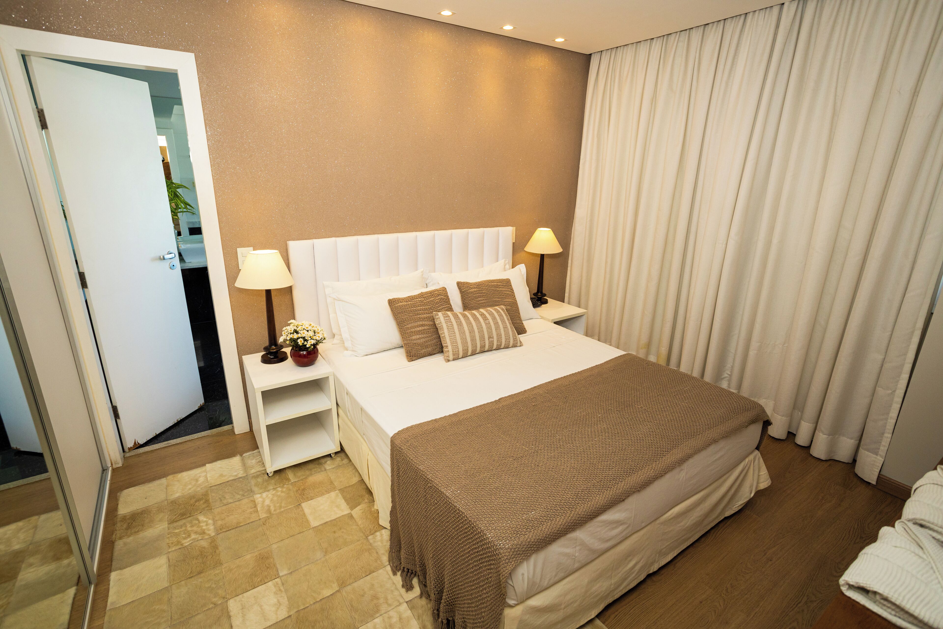 Quarto de hotel com uma cama grande, cabeceira branca, duas mesas de cabeceira com abajures, um cobertor marrom e vista para o corredor.