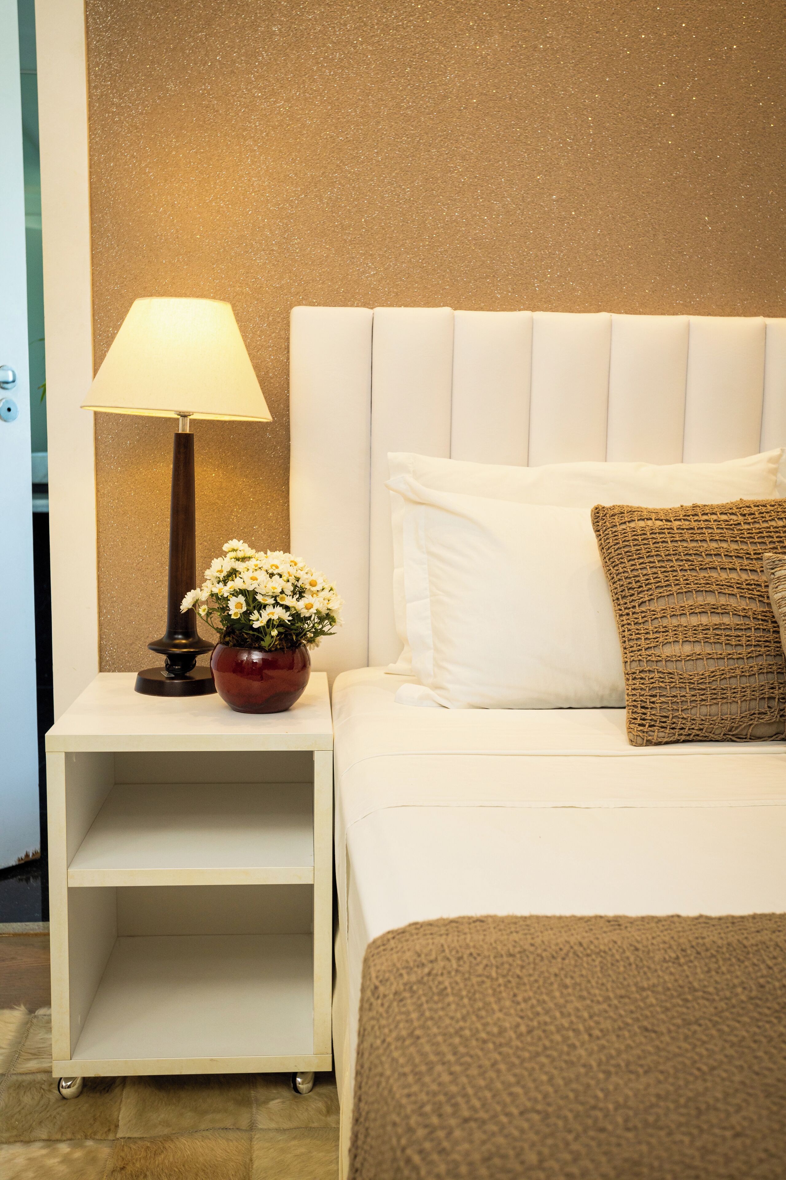 Un lit bien fait, avec une tête de lit blanche, une lampe de chevet, une petite table sur laquelle est posé un vase de fleurs et un coussin décoratif en tissu tissé.
