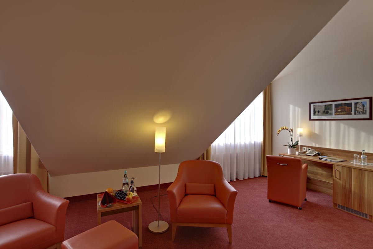 Ein Hotelzimmer mit schrägen Wänden, einem Schreibtisch mit Telefon, einem Sessel, einem kleinen Tisch mit Früchten und einer Stehlampe.