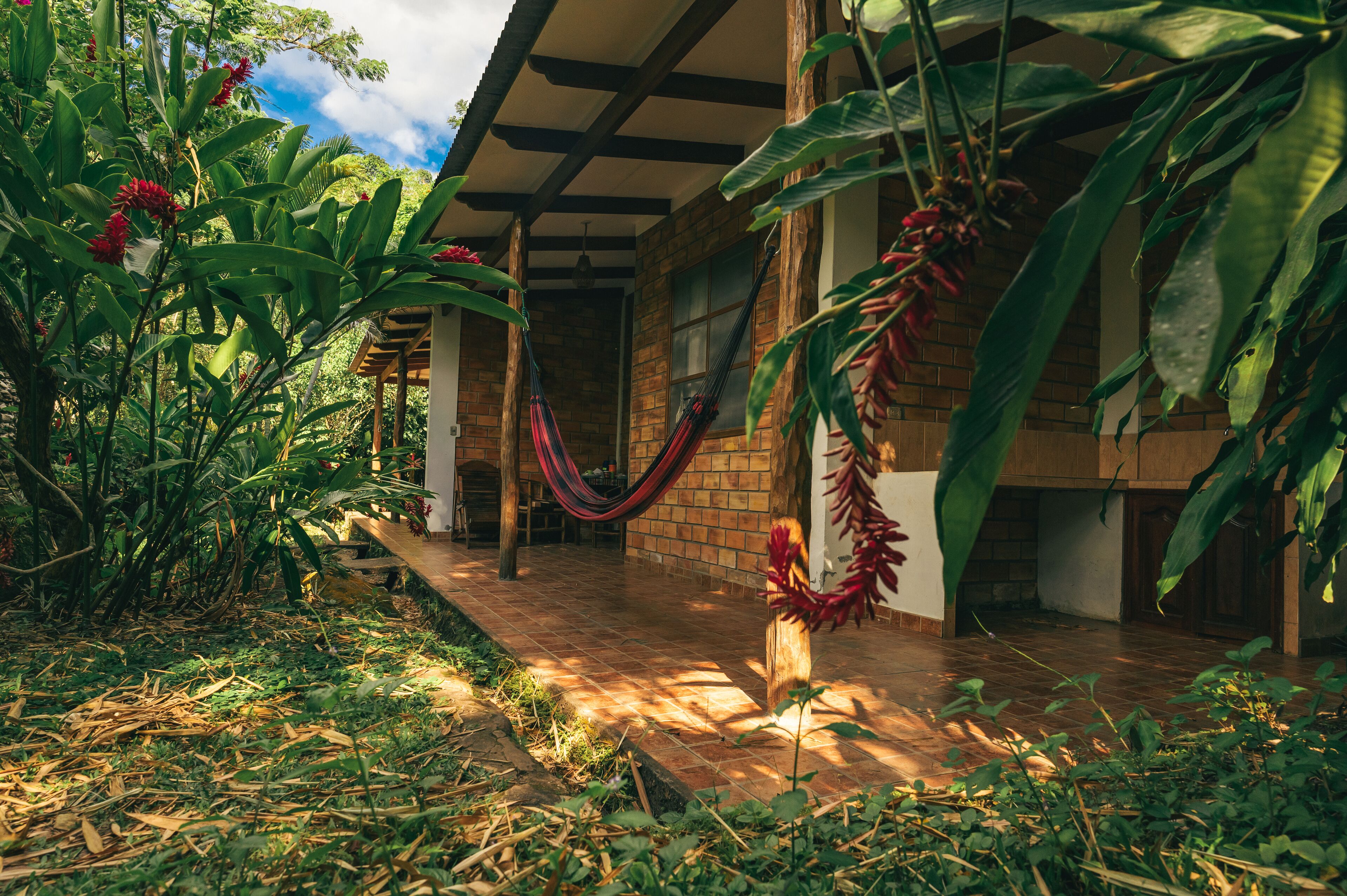 A covered outdoor area with a brick building, a hammock, and lush greenery.