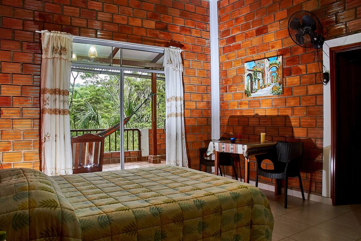 Quarto com cama, escrivaninha, cadeira, ventilador e janela com cortinas.