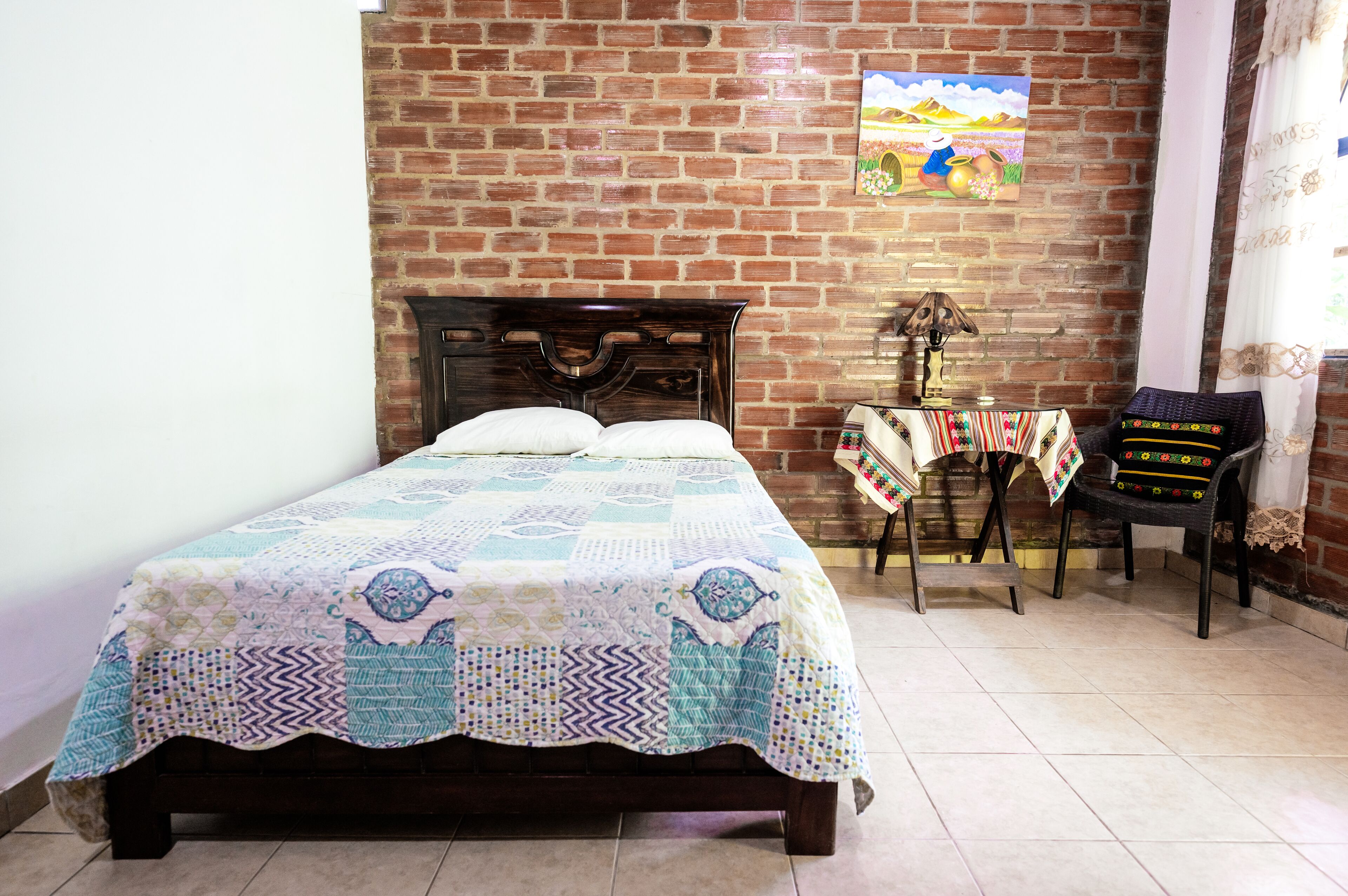 A room with a brick wall, a bed with a quilt, a small table with a lamp, and a chair with a colorful cushion.